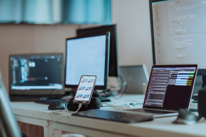 Blick auf mehrere Computer-Bildschirme und Smartphone auf einem Tisch mit laufenden Programmiercode in einem leeren Büro.