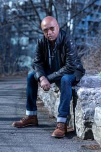 Jeff Jones is sitting on a cement wall wearing a leather jacket and sunglasses.