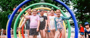 A group of young girls are playing in a water park, standing under colorful water arches and getting sprayed with water.