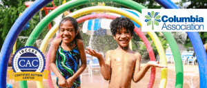 Two smiling children are playing in a water park, standing under colorful water arches and getting sprayed with water.