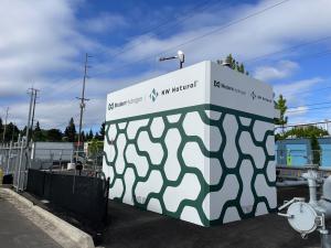 A hydrogen generation unit by Modern Hydrogen at NW Natural, featuring a white structure with green abstract patterns, located in a fenced area with urban surroundings in Portland, Oregon.