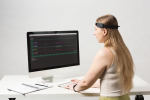 Woman working at a desk wearing a black headband. Caption: The new version of BrainAccess HALO is smaller, lighter and more comfortable. With low-energy Bluetooth connectivity (BLE), it can continuously stream EEG data for up to 9 hours.