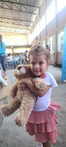 A female child holds a stuffed Phillip's Love Bears courage lion. Behind her are boxes of therapetuic stuffed animals and other children in seats with their own stuffed therapy animal..
