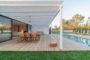 A wide-angle view of a modern villa featuring a swimming pool and a deck adorned with wooden furniture. The scene includes a partial interior view of the house, showcasing large glass doors seamlessly blending indoor and outdoor living spaces.