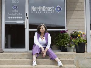 Owner and President of NorthCoast Media Group, LLC, sits outside the company's new location in Green Bay, Wisconsin. She is wearing a purple suit and sitting on the office steps, in the sun, with colorful flowers around her.