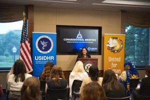 Latinex woman addresses a diverse group of women in front of large monitor reading "Congressional Briefing: Women in Diplomacy and Legislative Processes""