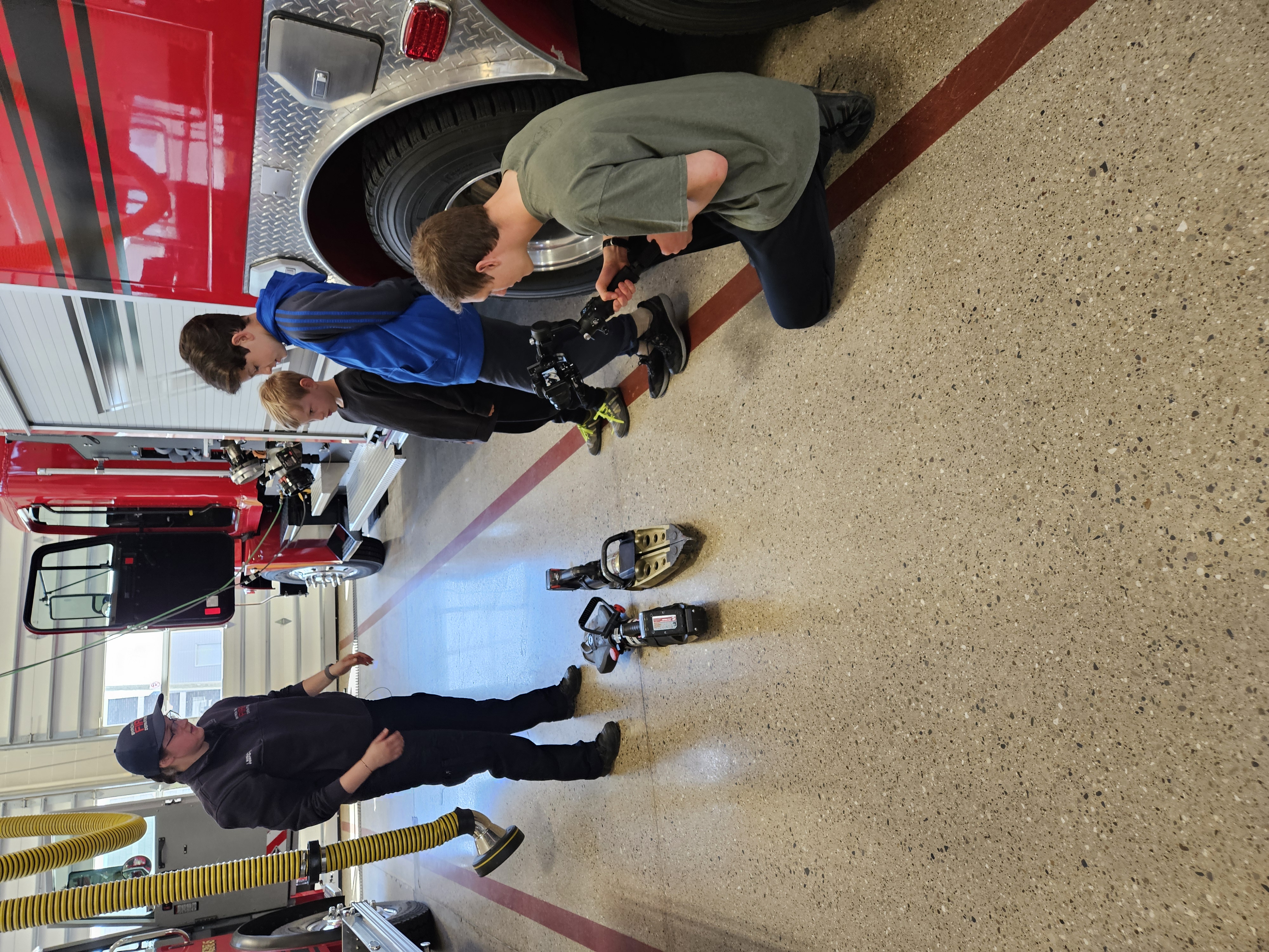 a firefighter gestures at cutters and spreaders, also known as the jaws of life, to show 2 boys how they work while being filmed for a #LifeSkillsNow workshop