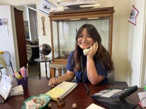 EXP summer intern Emily hard at work at the Los Angeles Maritime Institute with a phone to her ear and a pen in the other hand jotting notes. There are marketing materials scattered across the table.