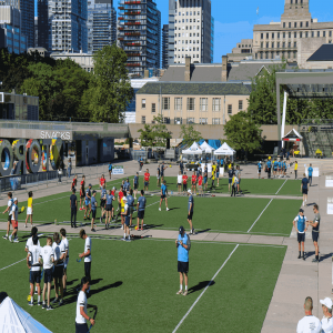  A wide view of the Rugby in the Square event at Nathan Phillips Square in Toronto. Multiple teams, dressed in different colored jerseys, are playing rugby on the green fields. The background features tall buildings and the iconic "Toronto" sign, with eve