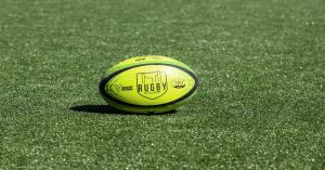 A close-up of a bright yellow rugby ball with the "Rugby in the Square" logo and Rugby Ontario branding, lying on the green artificial turf. The image captures the essence and excitement of the Rugby in the Square event at Nathan Phillips Square in Toront