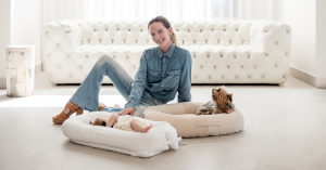 Woman with brown hair sits on floor in front of white couch. With her are a dog in a dog bed and a baby in a baby lounger.