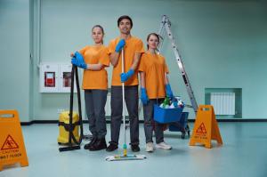 Three young workers of a cleaning company wearing orange shirts, gray pants, and blue latex gloves, posing with professional janitorial services tools, including a mop, vacuum cleaner, aluminum ladder, and disinfectant. A fire extinguisher and exhaust win