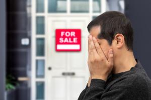 Extremely upset man with his head in his hands in front of a for sale sign on house