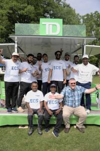 COTAPSA President Andrew Waters sitting with members of the City of Toronto ECS Cricket Team. The group is gathered in a tent outdoors, with in a TD Bank activation tent. Everyone is smiling and wearing cricket attire, showcasing a sense of camaraderie an