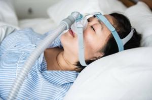 A woman with obstructive sleep apnea is sleeping on her white bed, wearing white and blue striped pajamas and an anti-snoring chin strap to help manage her condition.