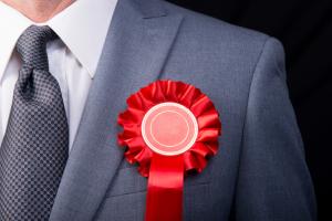 Grey suited businessman wearing a red rosette, against a black background