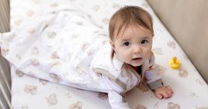 A baby lying on a patterned crib sheet, wearing a Burt's Bees Baby sleep sack, with a yellow pacifier nearby.