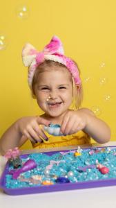Young girl enjoying countertop play with a blue splashy sand kit. The sensory tray contains mermaid themed imaginative play toys.