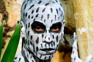 Itá Tupã dressed in black-and-white body paint with African-inspired designs, posing during the "Roots in Tupinambá Land 2022" photoshoot. The image highlights his deep and powerful expression, emphasizing a call to action for protecting indigenous lands.