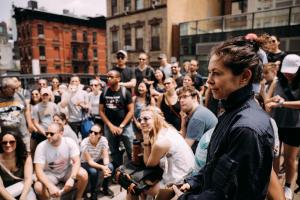 A Gotham Cluemaster with brown hair tied up, a smile on her face and dressed in blue coveralls oversees a large crowd of young New York event partcipants.