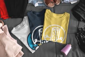 Three Dockhead t-shirts in white, navy blue, and yellow, featuring the Dockhead logo with a stylized dock and wave design. The shirts are neatly folded on a bed, surrounded by casual summer apparel, highlighting Dockhead’s eco-friendly and fashionable sum