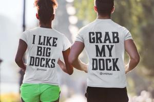 Two people wearing Dockhead t-shirts with humorous slogans. The woman’s shirt says ‘I Like Big Docks,’ and the man’s shirt says ‘Grab My Dock.’ They are walking together in an outdoor setting, showcasing Dockhead’s playful and bold apparel designed for su