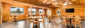 A warm, rustic dining hall with wooden walls, a vaulted ceiling, multiple tables and chairs, large windows, and a wall-mounted TV.
