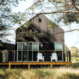 A cozy cabin with a wooden deck, two white chairs, and surrounded by tall trees and greenery.