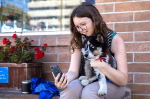A woman sits on a ledge outside at Berkeley Humane's Bark and Meow Around the Block event, smiling as she takes a selfie with her newly adopted black and white puppy with blue eyes. The puppy is wearing a red collar and appears calm and content.
