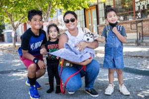 A smiling family poses outdoors with their newly adopted puppy at Berkeley Humane's Bark and Meow Around the Block event. The mother, holding the puppy wrapped in a blanket, is surrounded by her three young children, all looking happy and excited.