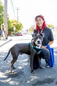 A proud adopter with red hair kneels next to their newly adopted gray and white dog wearing a teal harness at Berkeley Humane's Bark and Meow Around the Block event. They are outdoors on a sunny day, with a street and trees visible in the background.