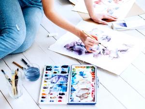 A lady uses water colors on papers spread out on the floor.