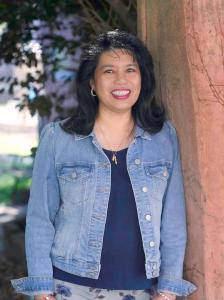 smiling tan-skinned woman with shoulder-length hair wearing a blue blouse and jean-jacket, standing outdoors and leaning on a brown stone wall