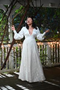 Mali Wilson sings passionately on a wooden veranda, wearing a long white gown, surrounded by greenery and twinkling lights, creating an enchanting atmosphere.