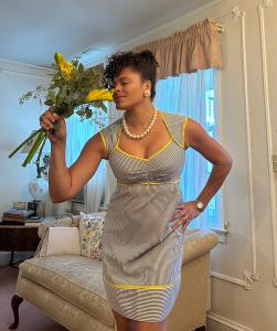 Mali Wilson stands in a cozy, elegantly furnished room, smiling as she smells a bouquet of yellow flowers while wearing a striped vintage-style dress with pearl jewelry.