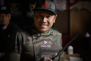  Chef Wijai “Song” Ketsuwan, dressed in a camo chef's jacket and hat, smiles while holding a knife at Sushi Song, showcasing his expertise and leadership in creating top-quality sushi in South Florida.