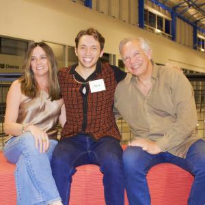 A smiling young man named Chase sits between two older adults, likely his parents, on a red bench in a brightly lit indoor setting. The woman on the left has long brown hair and is wearing a shiny sleeveless top and jeans. The man on the right has gray ha