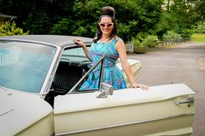 Mali Wilson channels retro elegance in a vintage-inspired dress and sunglasses, posing beside a classic car, reflecting her unique style and creative vision.