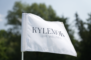 A close-up image of a white pin flag from the 2024 Markham Board of Trade Corporate Charity Golf Classic, featuring the title sponsor's logo, "KYLEMORE." The flag displays the words "KYLEMORE" in bold black letters with the tagline "LIVE. WELLNESS." below