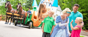 A family smiles and interacts with a princess character near a horse-drawn carriage in front of a fairy tale castle.