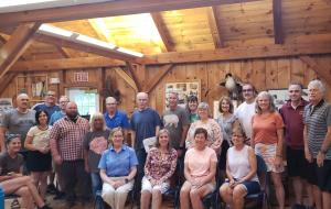 A group of 21 people gather for a photo inside what looks to be a wooden structure. It is a mix of men and women.