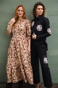 Two women wearing Transcend's Empower Collection Pieces - the woman on the right is wearing the black Jordan jacket, black Riley pants and the woman on the left is wearing a brown and beige print maxi dress. Both women are leaning against an olive green w