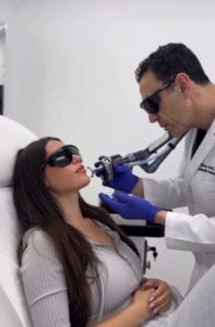 r. Simon Ourian performs a Coolaser treatment on Yolanthe Cabau, who is lying on a treatment bed wearing protective eyewear, while Dr. Ourian uses a precision laser device.