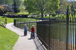 Two young boys riding bicycles stop around a duck pond in a park enclosed by a black steel fence, with trees and greenery in the background.