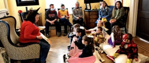 Several children are seated on the floor of a living room, listening to woman's storytelling.