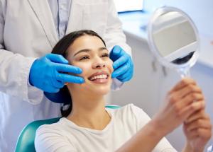 A young woman smiles while examining her teeth in a handheld mirror after a dental procedure. The dentist's hand in blue gloves gently positions her face to showcase the transformation.