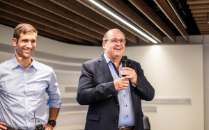 Adrian Brüngger und Hansruedi Wipf präsentieren auf einer Konferenz. Brüngger trägt ein hellblaues Hemd mit Headset, während Wipf, gekleidet in einen dunklen Anzug, ein Mikrofon in der Hand hält. Beide lächeln