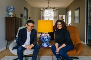 Amitābha Studio founders Davide Piai and Kimberly Poppe seated in a stylish living room, with a blue and white lamp from their Royal Delft collaboration, featuring a yellow shade, on a table between them. The room showcases modern furniture and decor with