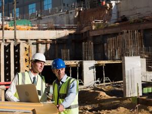 Two male engineers in safety gear oversee a construction site, reviewing project details on a laptop on a protective, glossy, plastic-covered surface.