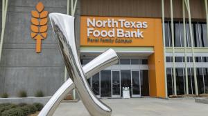 Exterior view of the North Texas Food Bank Perot Family Campus featuring a large orange logo resembling a wheat sheaf beside a shiny, metallic sculpture in the foreground, under a clear sky.